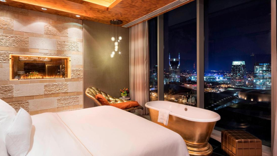 Luxurious hotel room with a large bed, golden freestanding bathtub, and floor-to-ceiling windows offering a nighttime city view. The room features elegant lighting, a modern fireplace, and plush seating