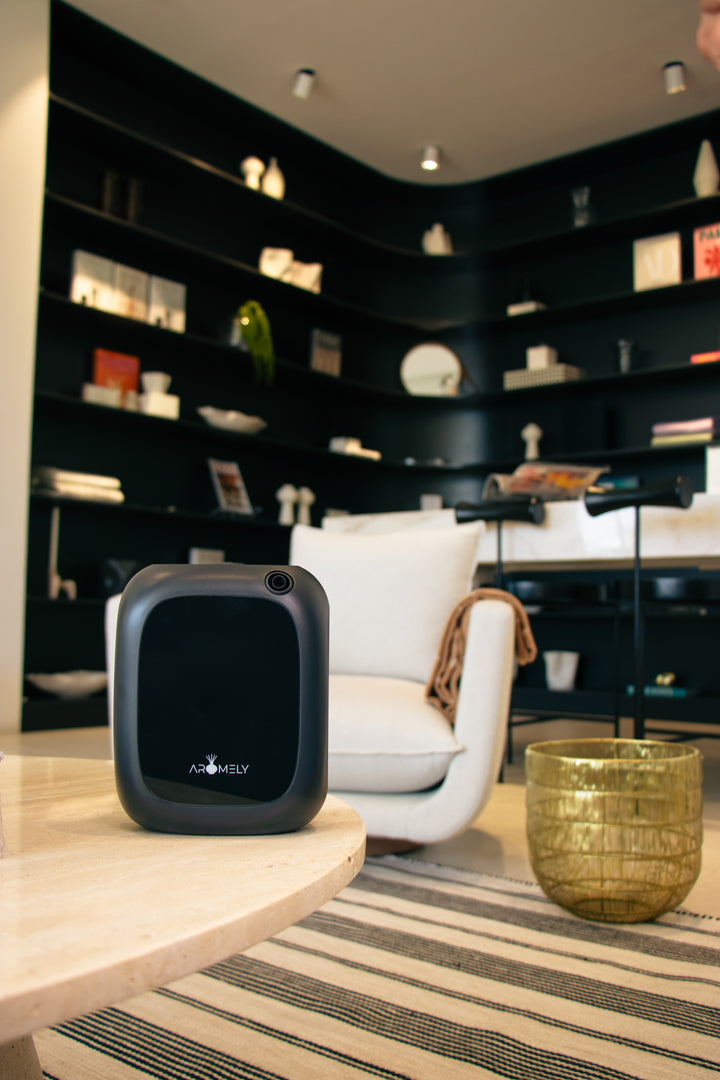 "A contemporary room with a black Aromely ARO-25-MAX scent diffuser on a light wooden table. The diffuser contrasts with the white armchair and golden decorative bowl, set against a backdrop of stylish black bookshelves with various decorative objects.