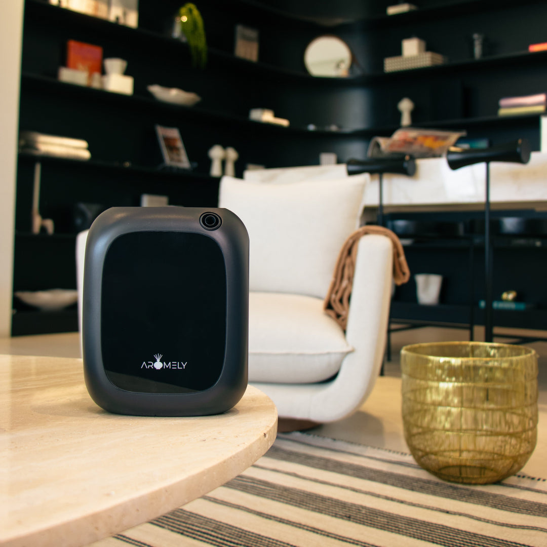 "A contemporary room with a black Aromely ARO-25-MAX scent diffuser on a light wooden table. The diffuser contrasts with the white armchair and golden decorative bowl, set against a backdrop of stylish black bookshelves with various decorative objects.