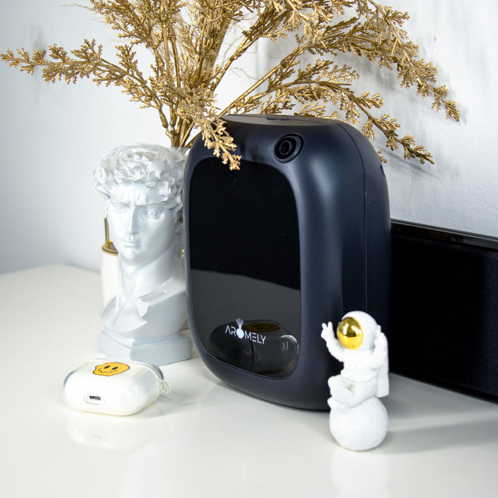 A chic and modern bathroom setting showcasing a black Aromely ARO-25-MAX scent diffuser on a white countertop. The diffuser is accompanied by artistic decorations including a golden spray of dried plants and a classical bust, creating a luxurious ambiance