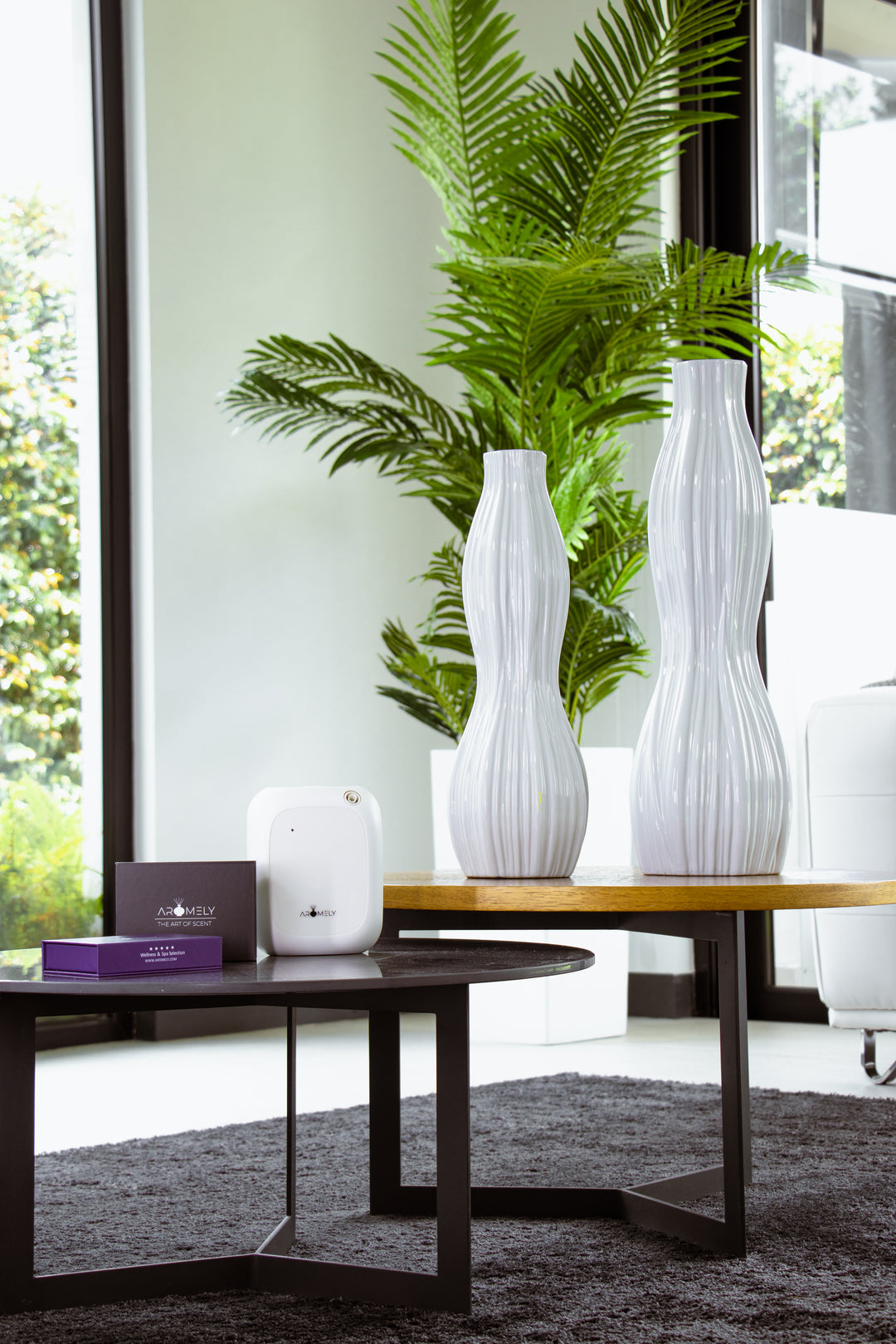 A modern and minimalist living room featuring an Aromely ARO-25-MAX white scent diffuser on a wooden console table. The diffuser is placed next to sleek white vases and a lush green potted plant, with a large window providing natural light.