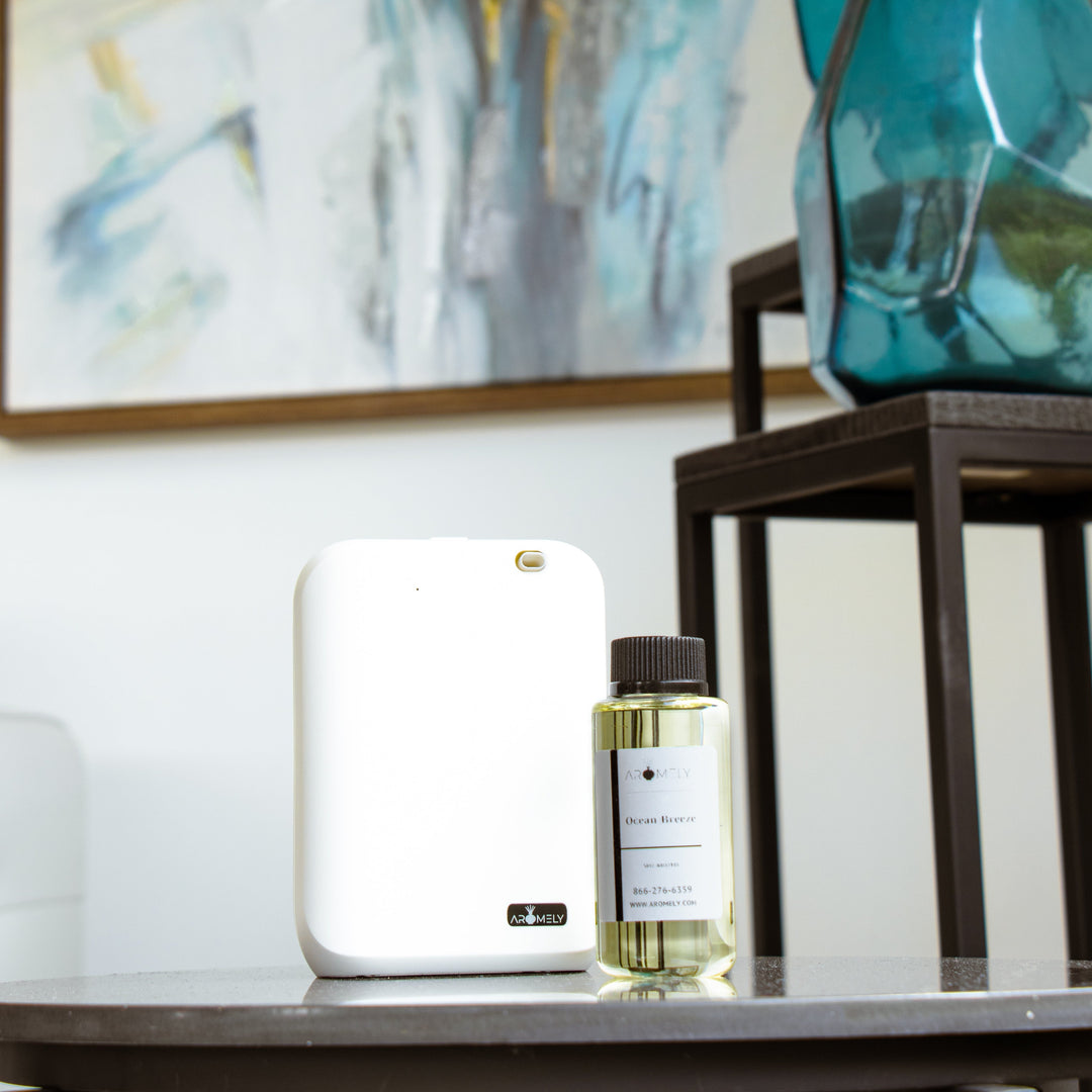 Aromely ARO-X25 Smart Aroma Diffuser placed on a table with a bottle of essential oil, with abstract art and a decorative blue vase in the background.