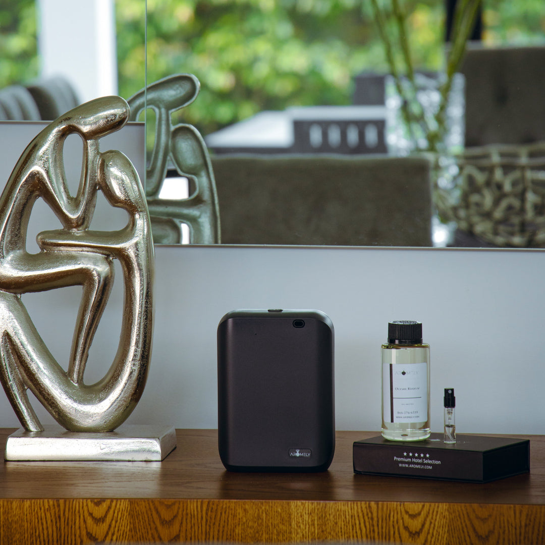Aromely ARO-X25 Smart Aroma Diffuser in a dark color, placed on a wooden shelf with a modern art sculpture and fragrance bottle.