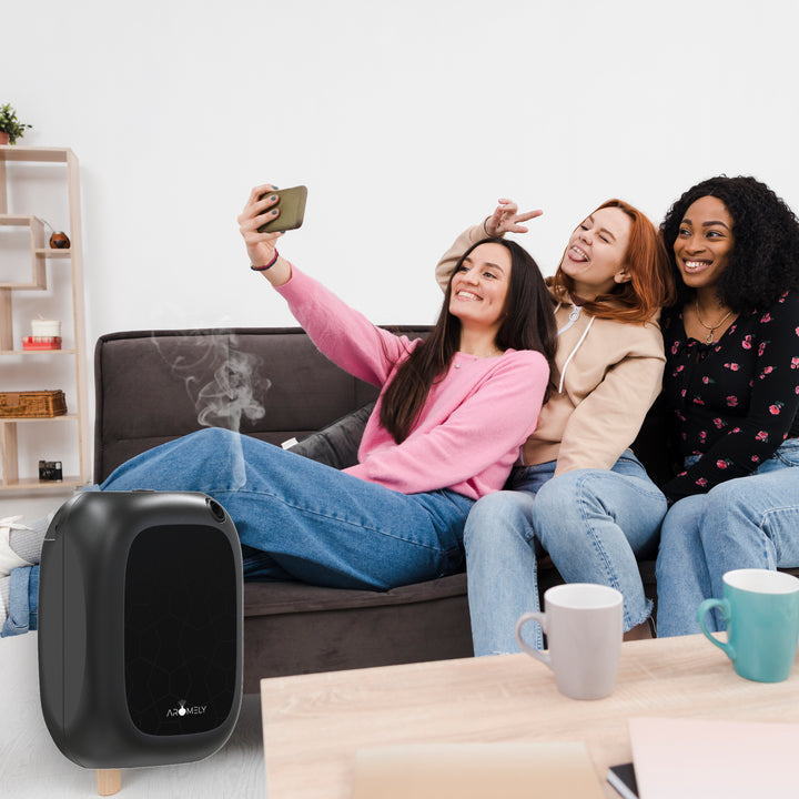 A lighthearted moment captured as three friends take a selfie on a sofa, with the Aromely ARO-25-MAX black scent diffuser on a low coffee table, subtly releasing scented vapor, contributing to the cheerful ambiance in the bright, modern living room