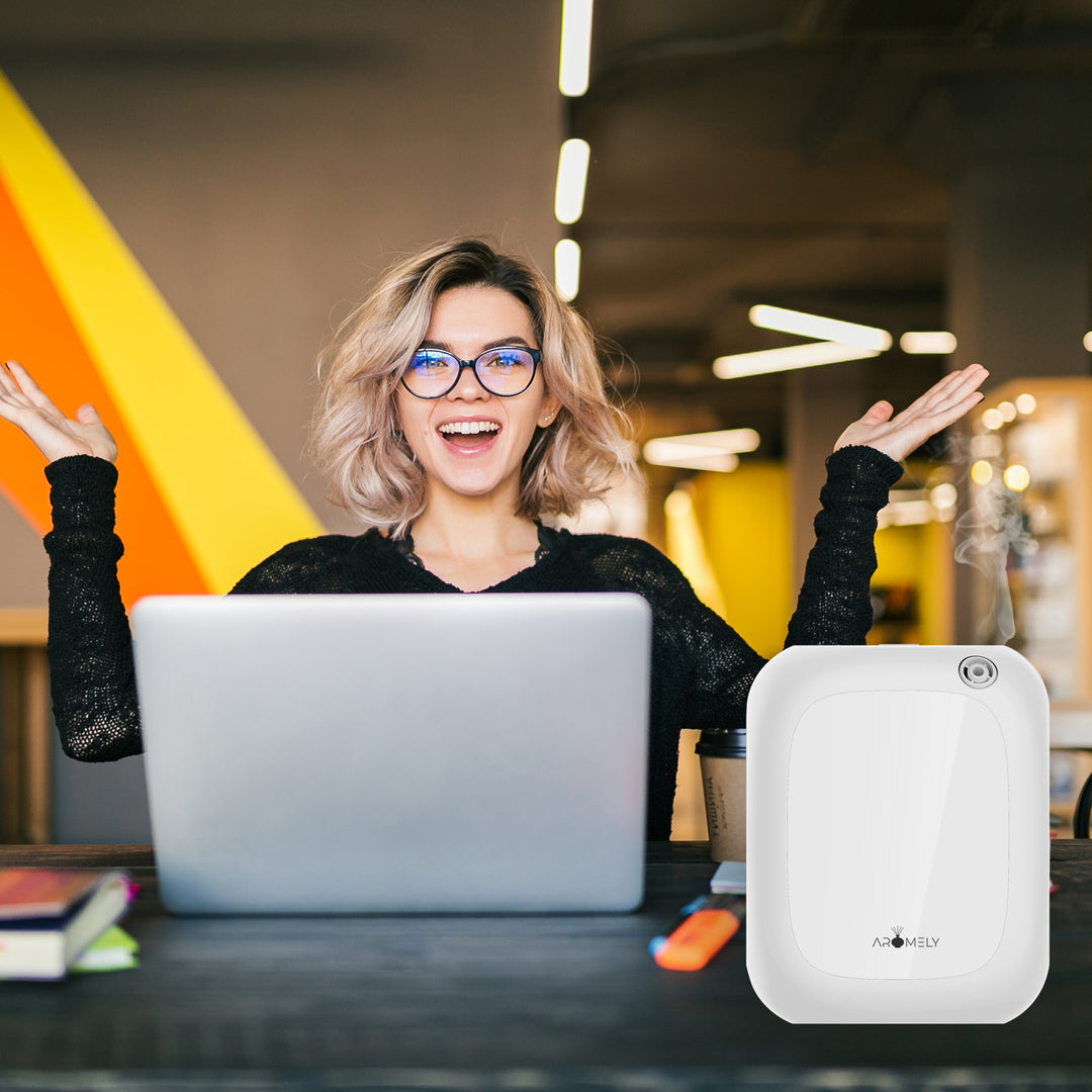 A lively atmosphere in a creative workspace, where a young woman with short, wavy hair expresses surprise and joy, sitting at a desk with the Aromely ARO-25-MAX white scent diffuser nearby, which adds to the dynamic and inspirational environment.
