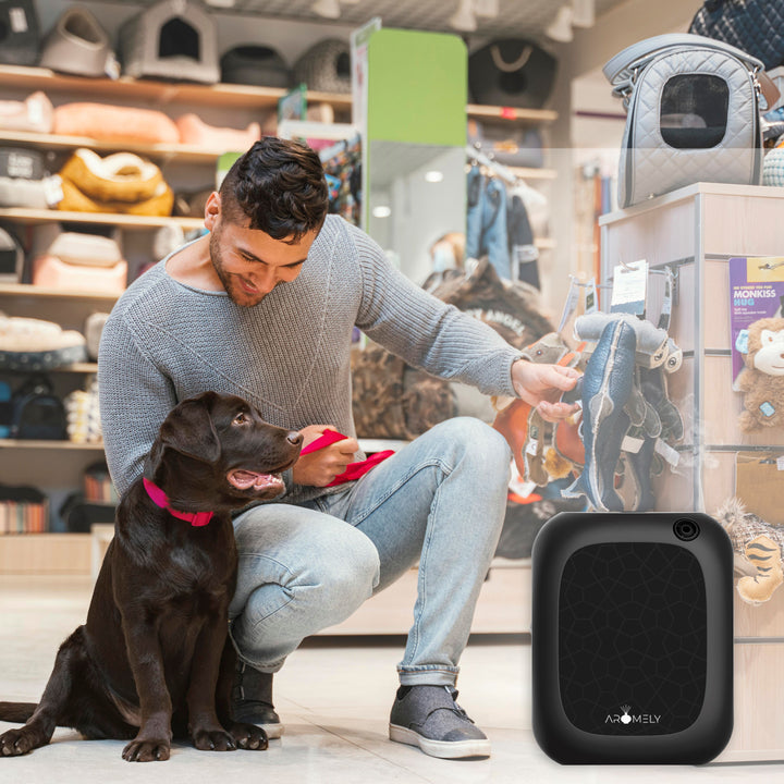 A heartwarming scene in a pet store where a man crouches to show a brown dog a new toy, with a black Aromely ARO-25-MAX scent diffuser in the foreground. The diffuser enhances the shopping experience with its calming scent amidst the colorful array of pet supplies