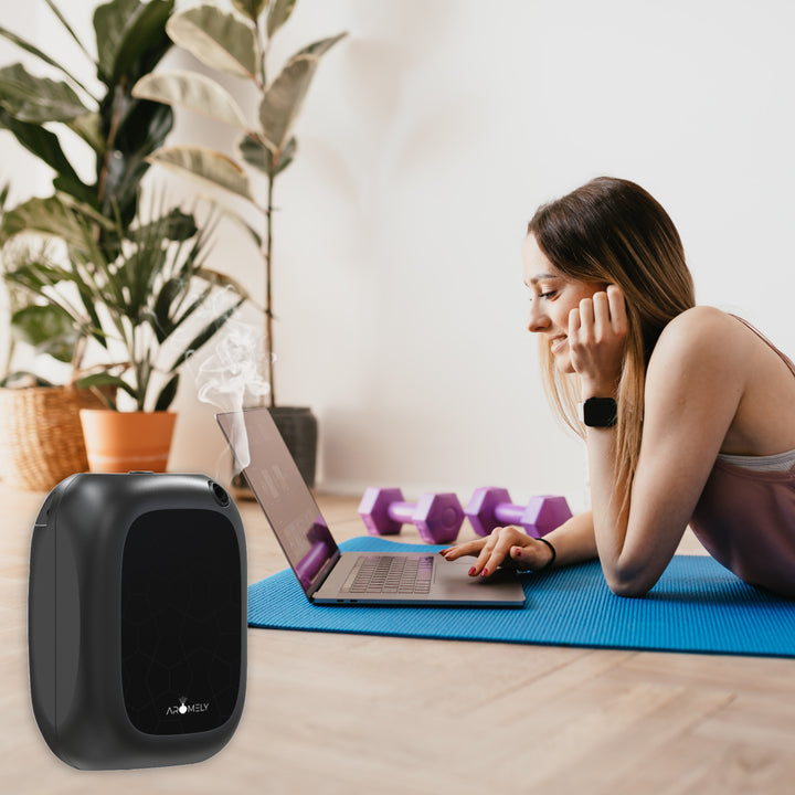 An active setting with a focused woman lying on a yoga mat, using a laptop next to a black Aromely ARO-25-MAX scent diffuser. The diffuser sits on the floor releasing a relaxing fragrance, complementing the peaceful indoor workout space adorned with potted plants.